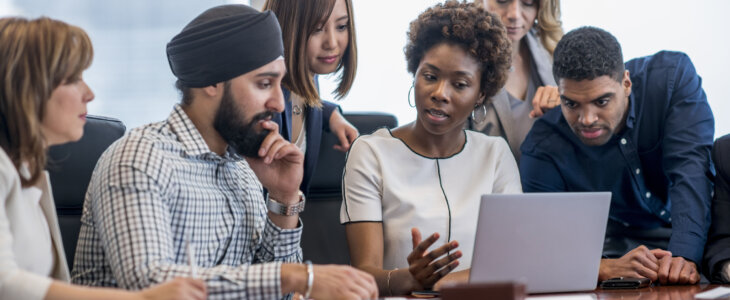 A diverse group of business people gather around a laptop in a modern office and discuss what they see.